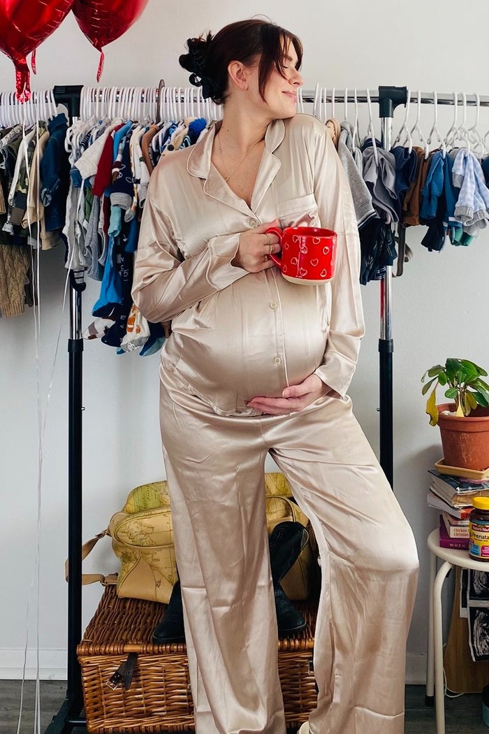 woman wearing a beige silky maternity pajama set holding a red mug and showing her baby bump