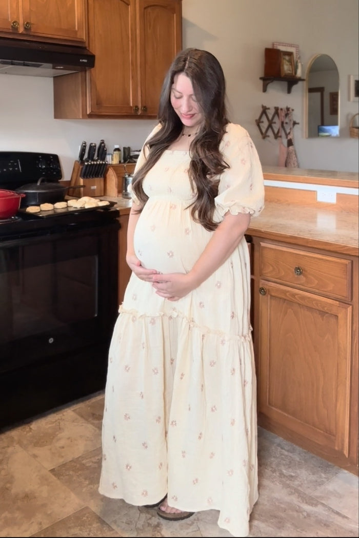 woman wearing a light yellow floral maternity maxi dress and holding her baby bump in a kitchen setting