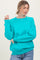 woman wearing a turquoise oversized sweatshirt and light blue jeans, smiling with a playful pose