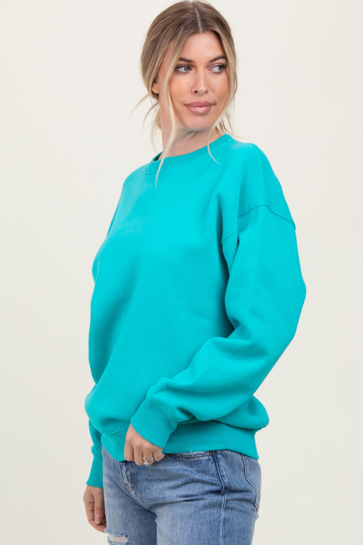 woman wearing a bright turquoise oversized sweatshirt and blue jeans, standing with a slight smile