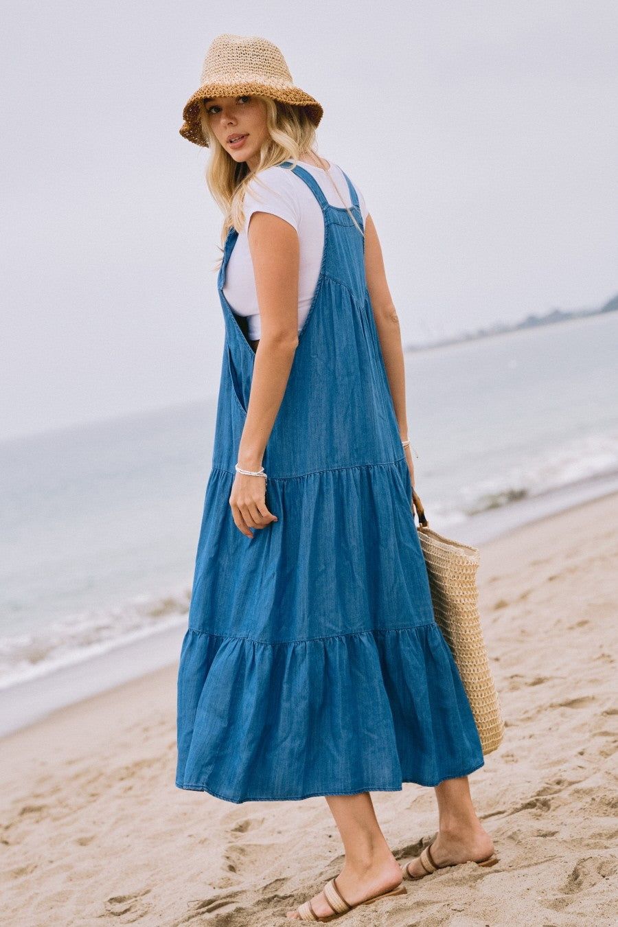 woman wearing a blue denim tiered maxi dress with a white t-shirt underneath and a straw hat by the beach