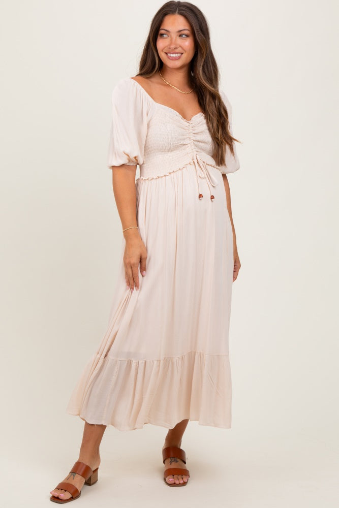 woman wearing a light beige maternity dress with a ruched bodice and puff sleeves, standing against a neutral background