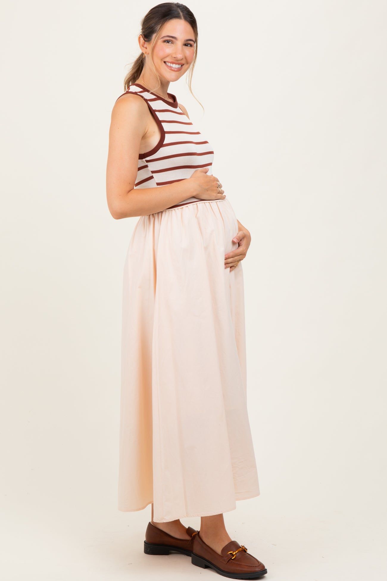woman wearing a striped sleeveless maternity top and a long peach skirt, holding her baby bump