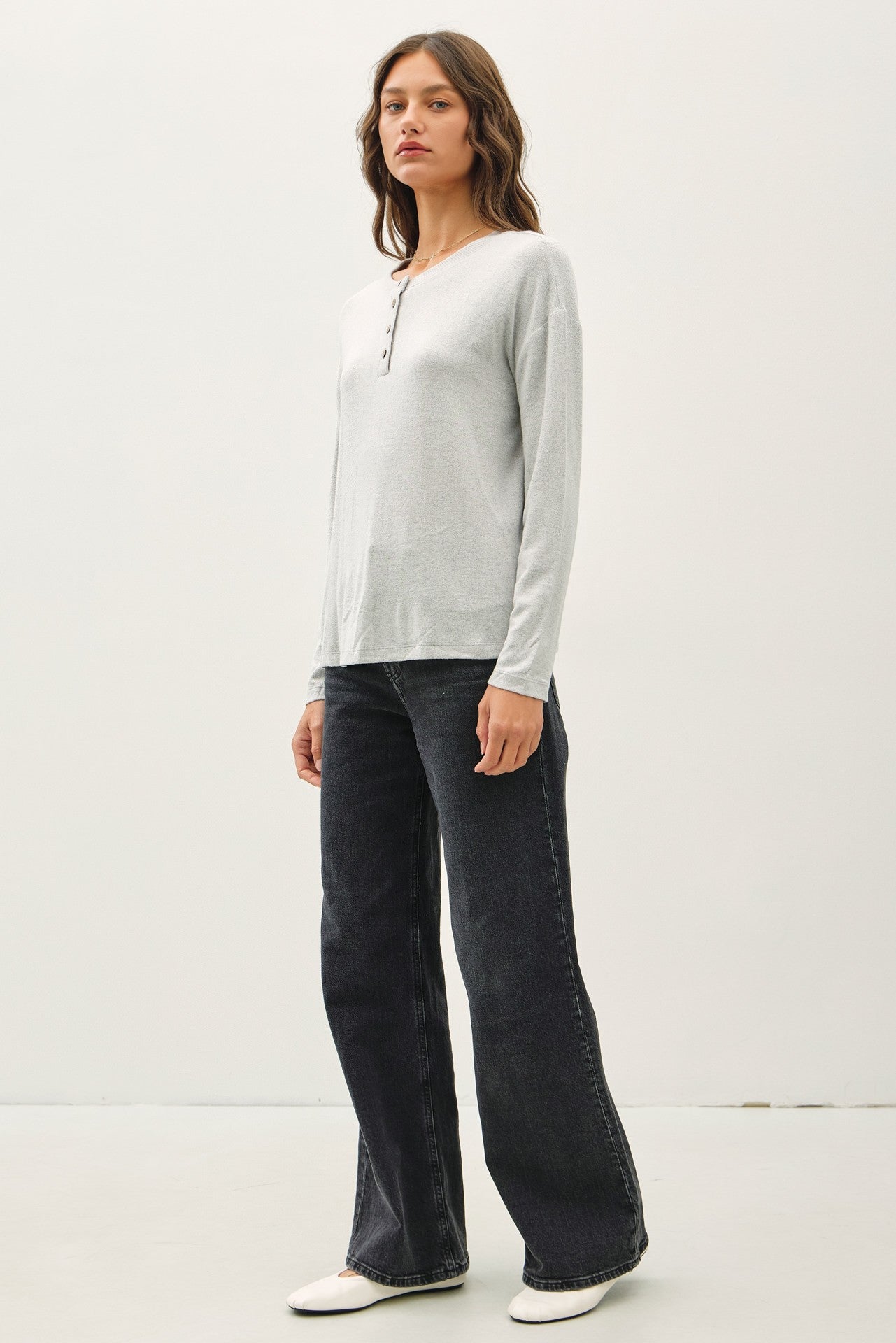 woman wearing a light gray long sleeve henley top and dark wide-leg jeans, standing against a plain background