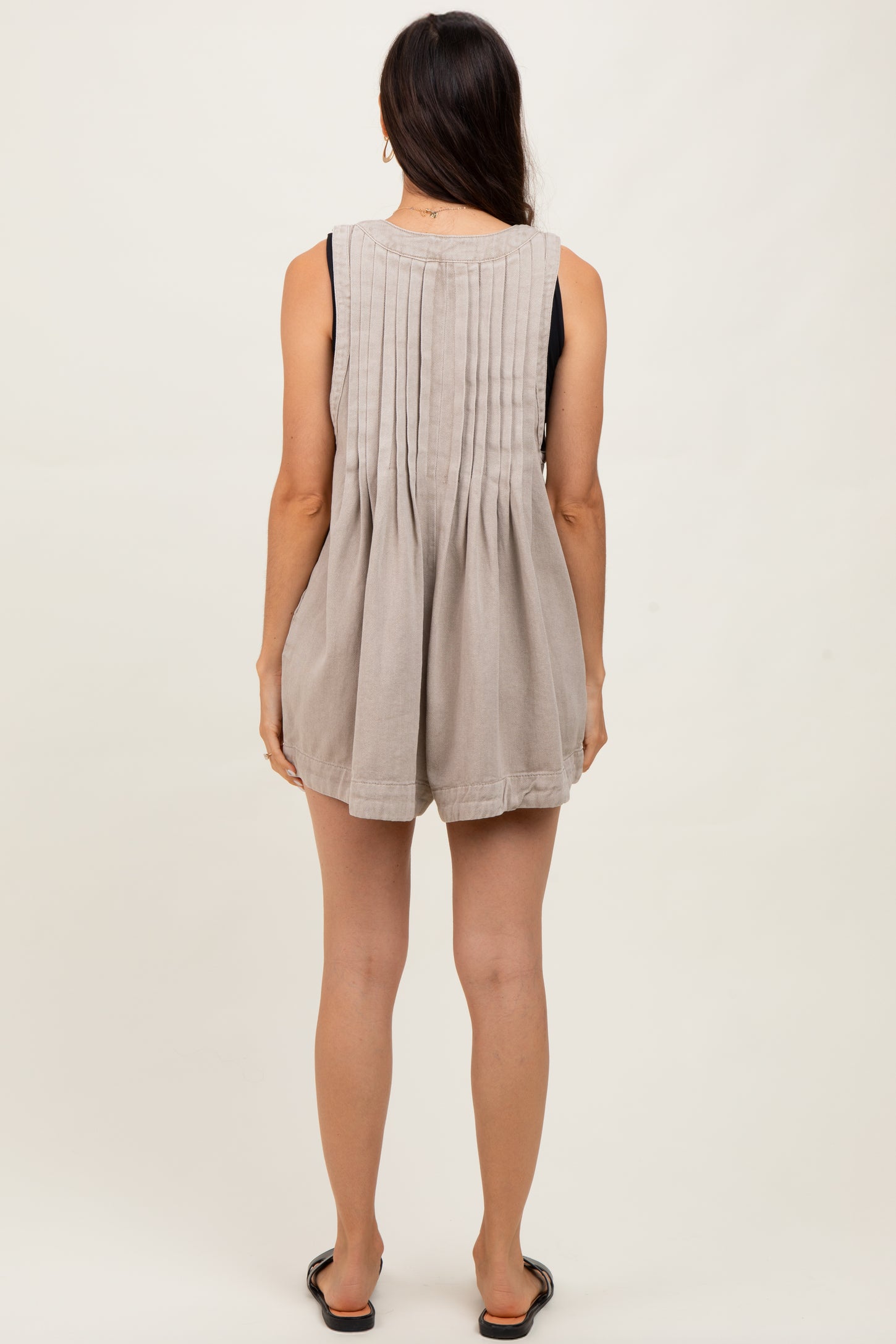 woman wearing a beige sleeveless romper with a pleated back detail, viewed from the back