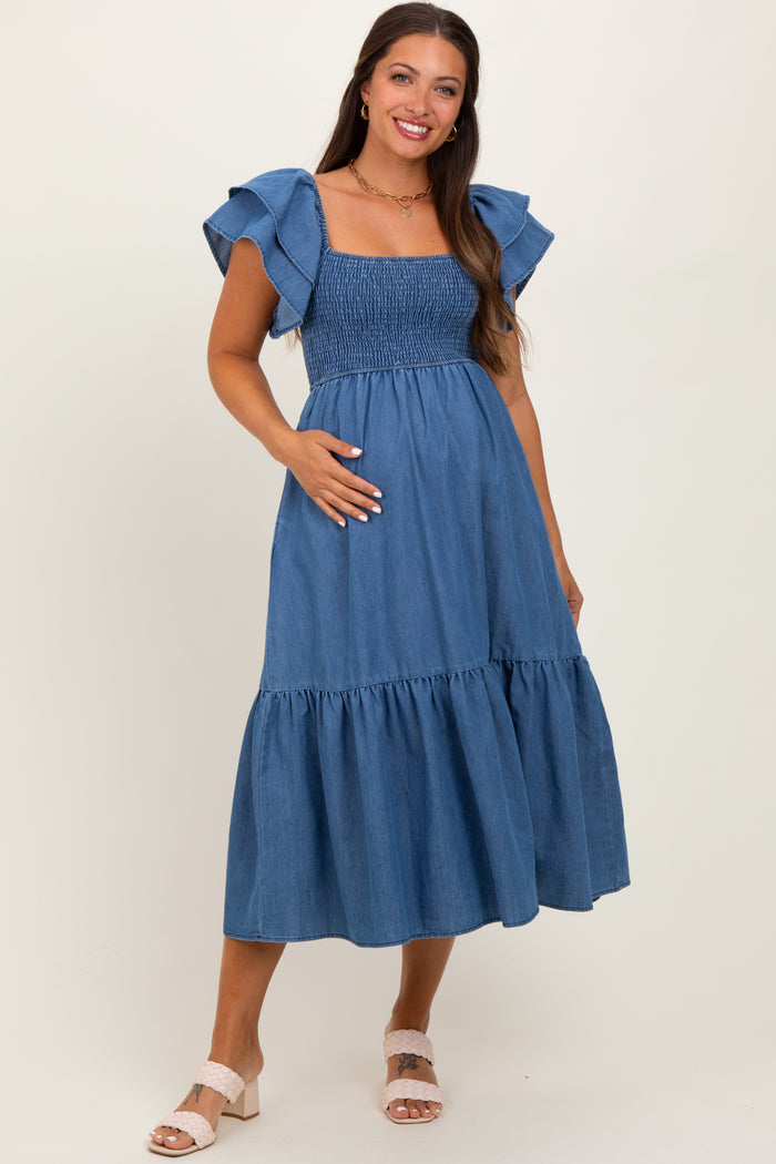 woman wearing a blue denim maternity dress with ruffled sleeves and a smocked bodice, holding her baby bump