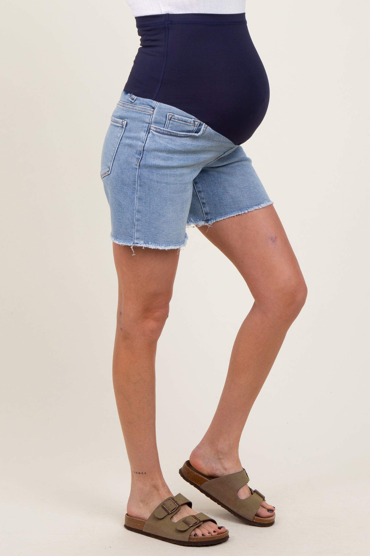 woman wearing distressed light wash maternity shorts with a dark blue belly band and brown slip-on sandals, showing baby bump