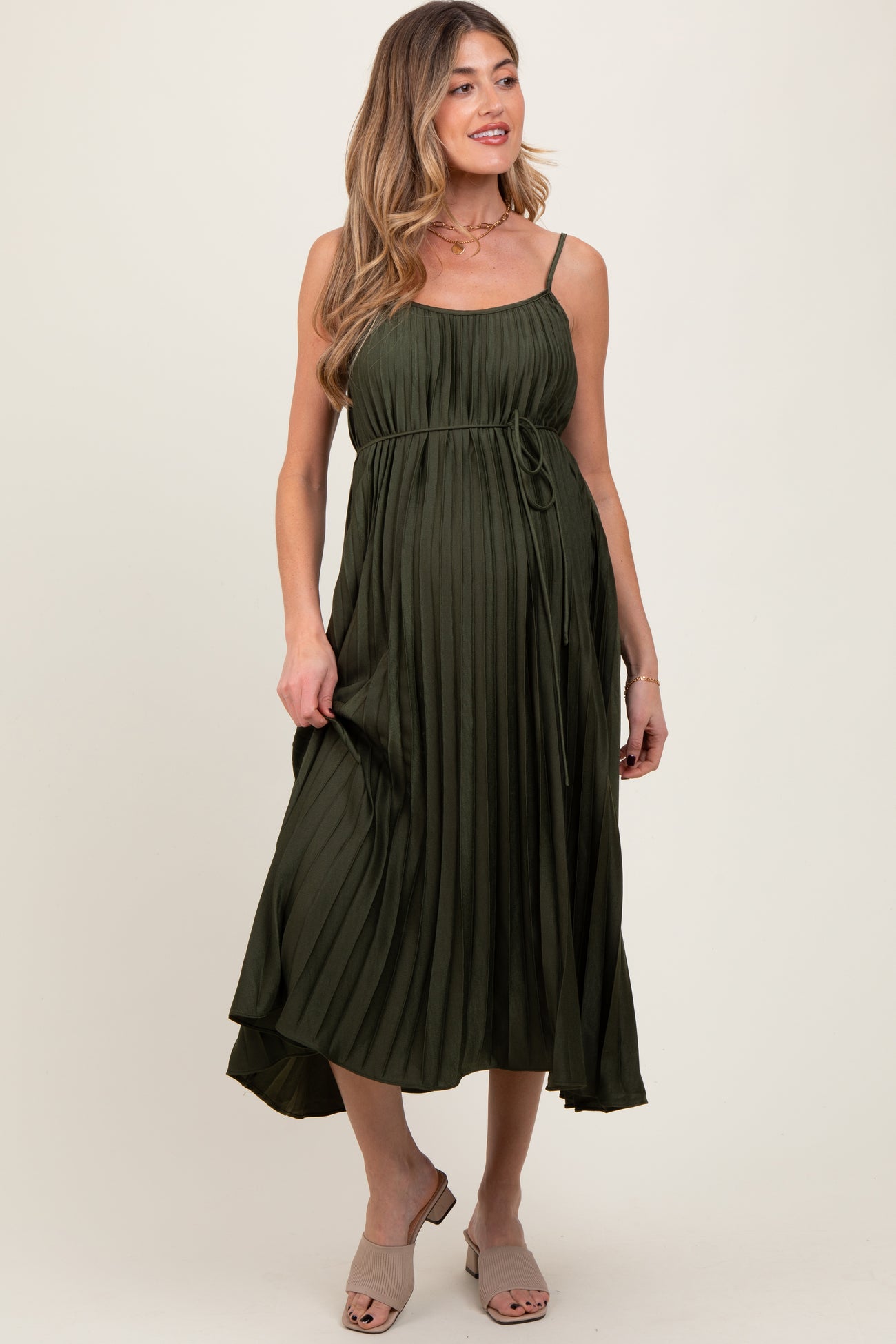 woman wearing an olive green pleated maternity dress with a tie waist and adjustable spaghetti straps