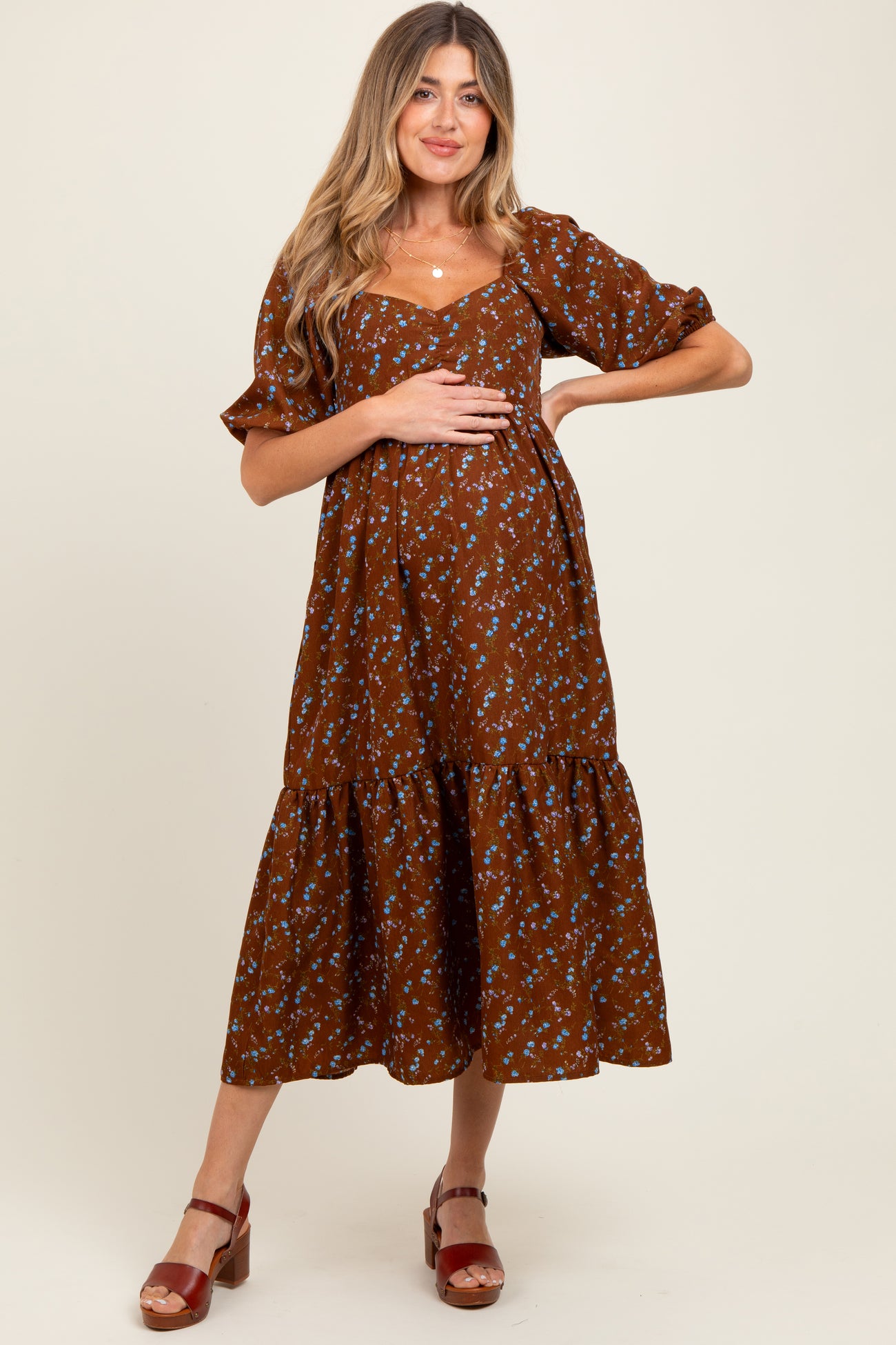 woman wearing a brown floral maternity dress with puff sleeves and tiered skirt, gently holding her baby bump