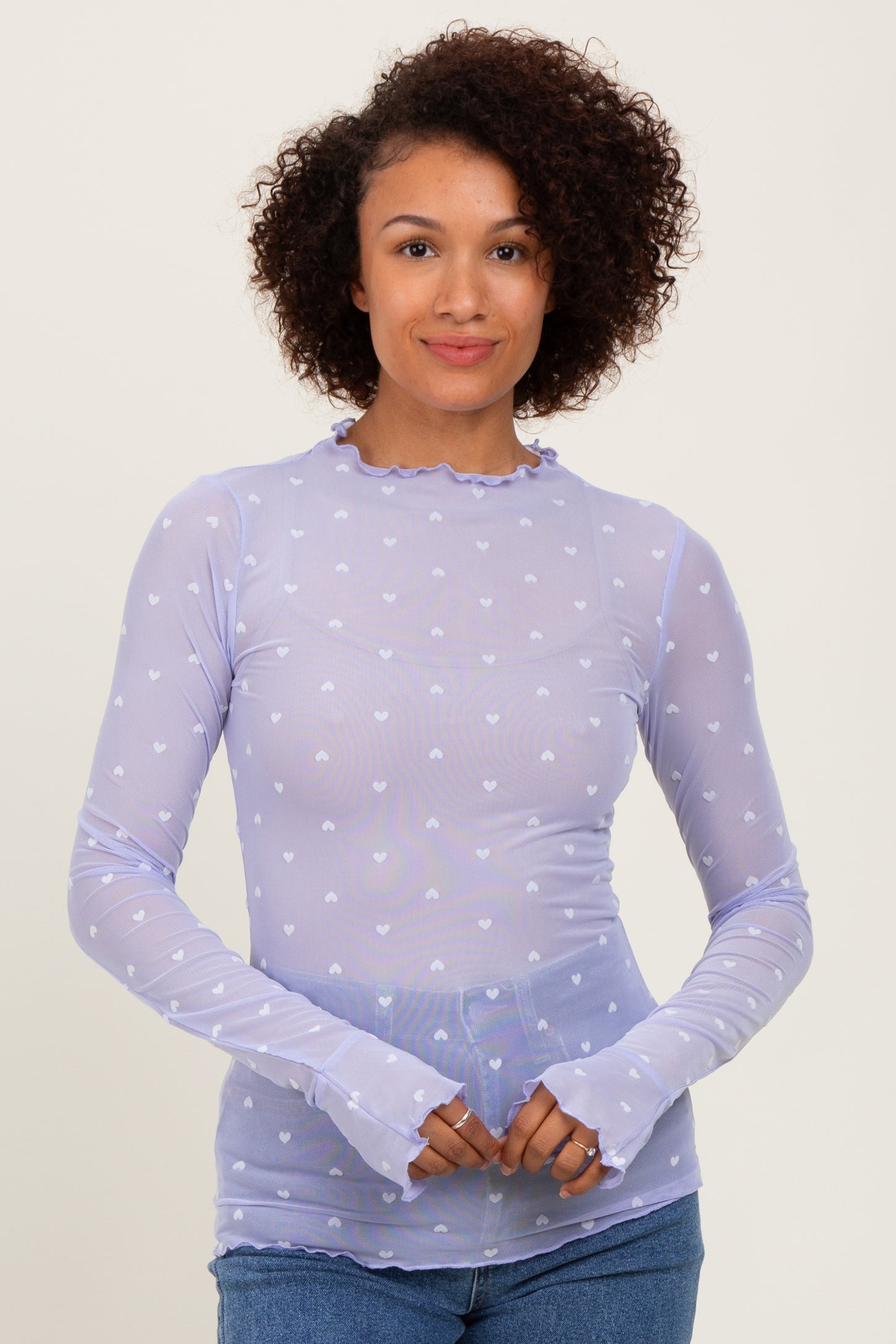 woman wearing a lavender long sleeve sheer top with heart patterns and ruffled neckline, posing with hands together