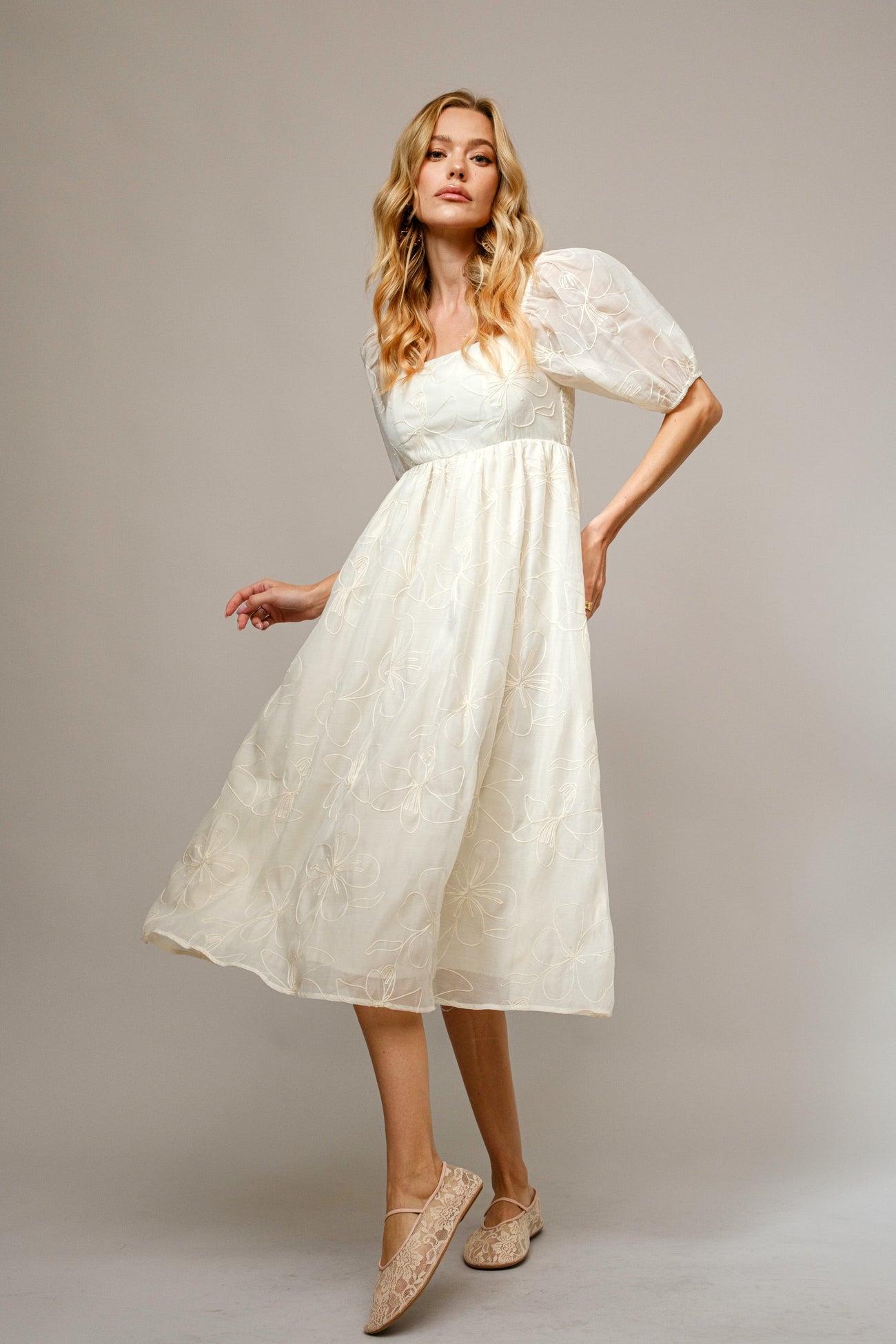 woman wearing a cream mid-length puff sleeve dress with floral embroidery and beige flats
