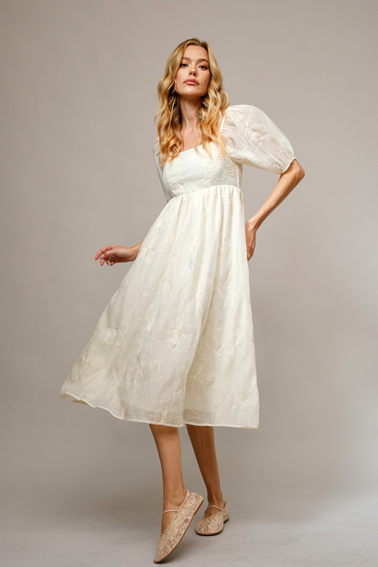 woman wearing a cream mid-length puff sleeve dress with floral embroidery and beige flats