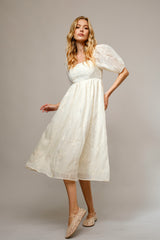 woman wearing a cream mid-length puff sleeve dress with floral embroidery and beige flats