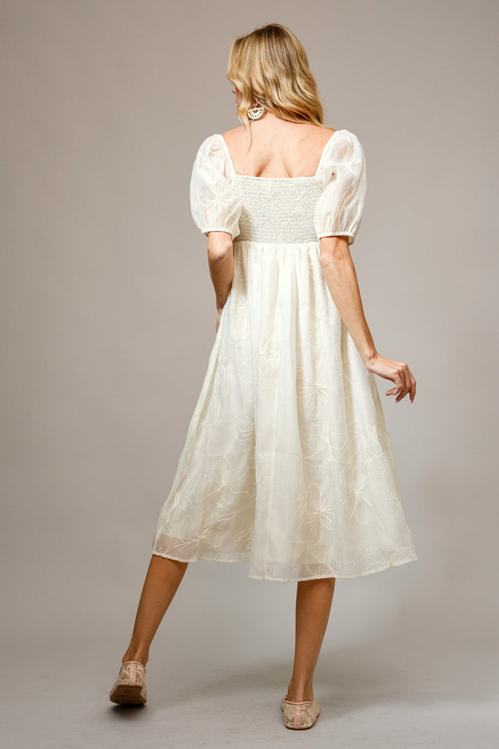 woman wearing a cream-colored midi dress with puff sleeves and a smocked bodice, standing with her back to the camera