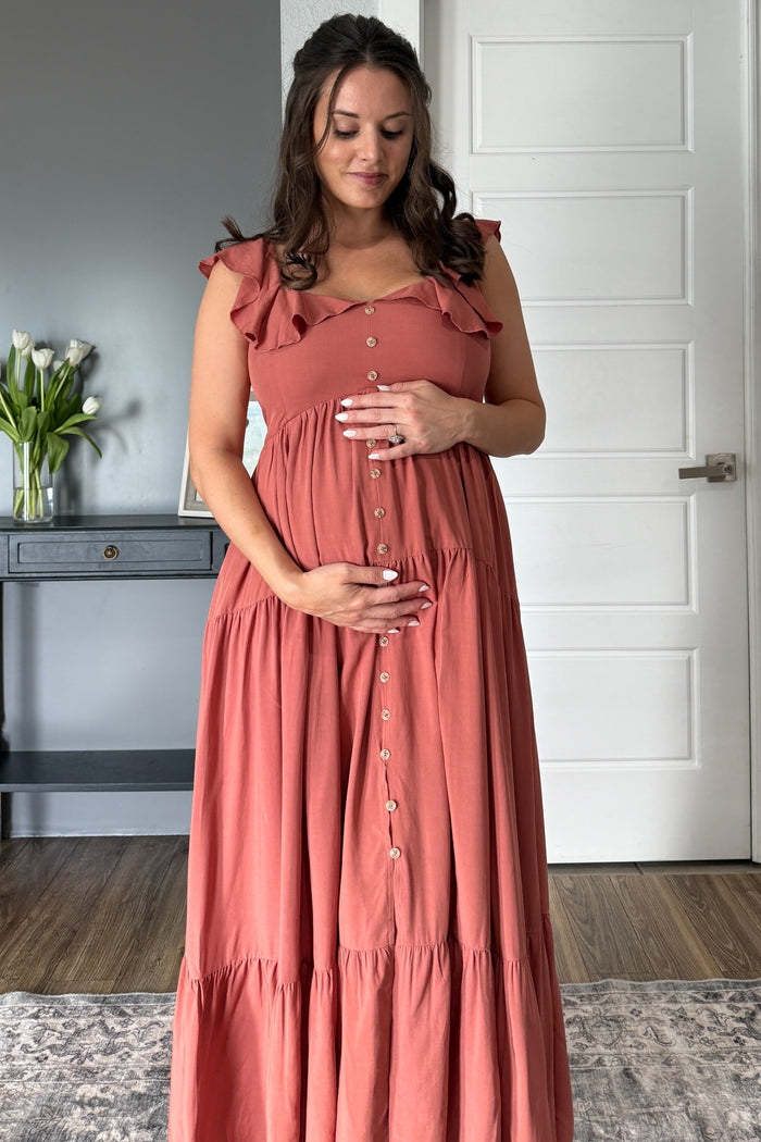 woman wearing a rust-colored maternity maxi dress with ruffle straps and a button-down front, holding her baby bump