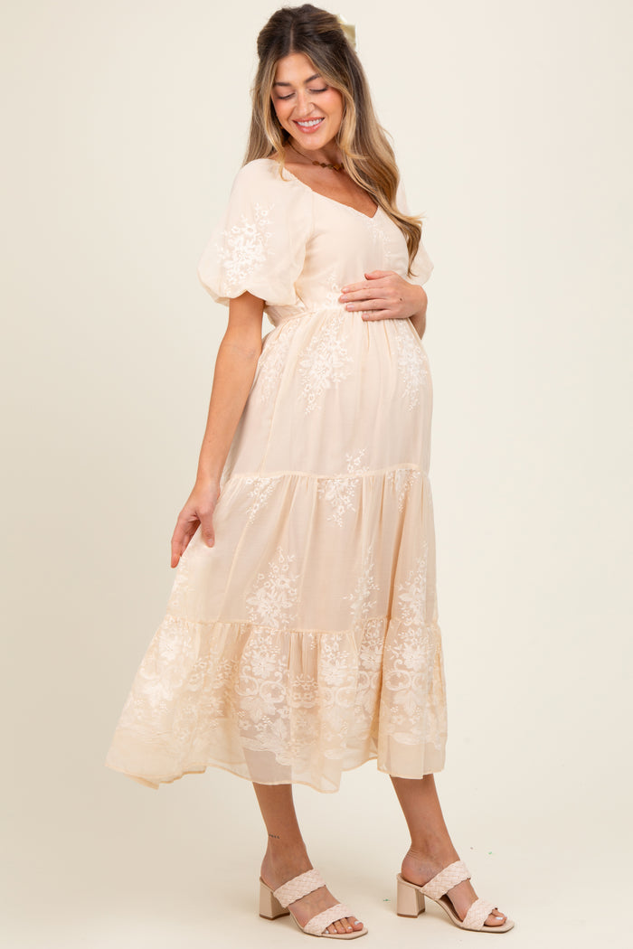 woman wearing a light peach maternity dress with floral lace details and a flowing skirt, cradling her baby bump
