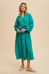 woman wearing a teal long sleeve button-up dress with a tie waist and a flowing skirt, standing on a beige background