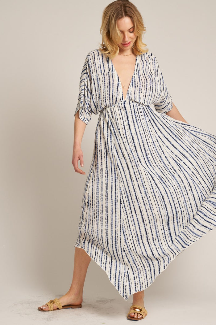 woman wearing a flowy white maxi dress with navy stripes and a deep v-neck, standing in a playful pose