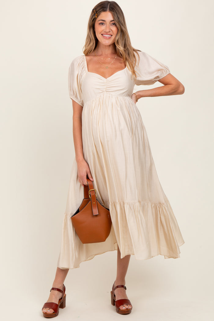woman wearing a beige maternity dress with puff sleeves and a tiered hem, holding a brown handbag and showing her baby bump