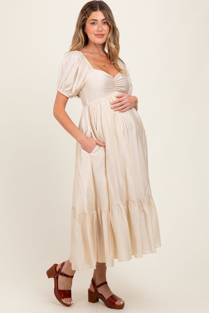 woman wearing a beige maternity dress with puff sleeves and front ruching, holding her baby bump and standing in a relaxed pose