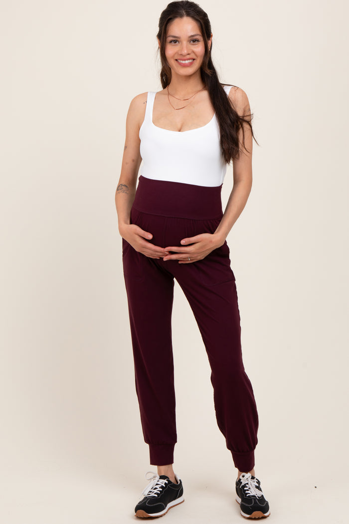 woman wearing a white maternity bodysuit and maroon maternity joggers, gently holding her baby bump