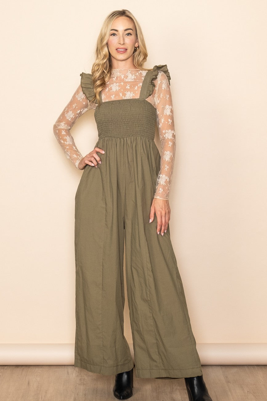 woman wearing an olive green jumpsuit with ruffled straps over a sheer floral long sleeve top and black ankle boots