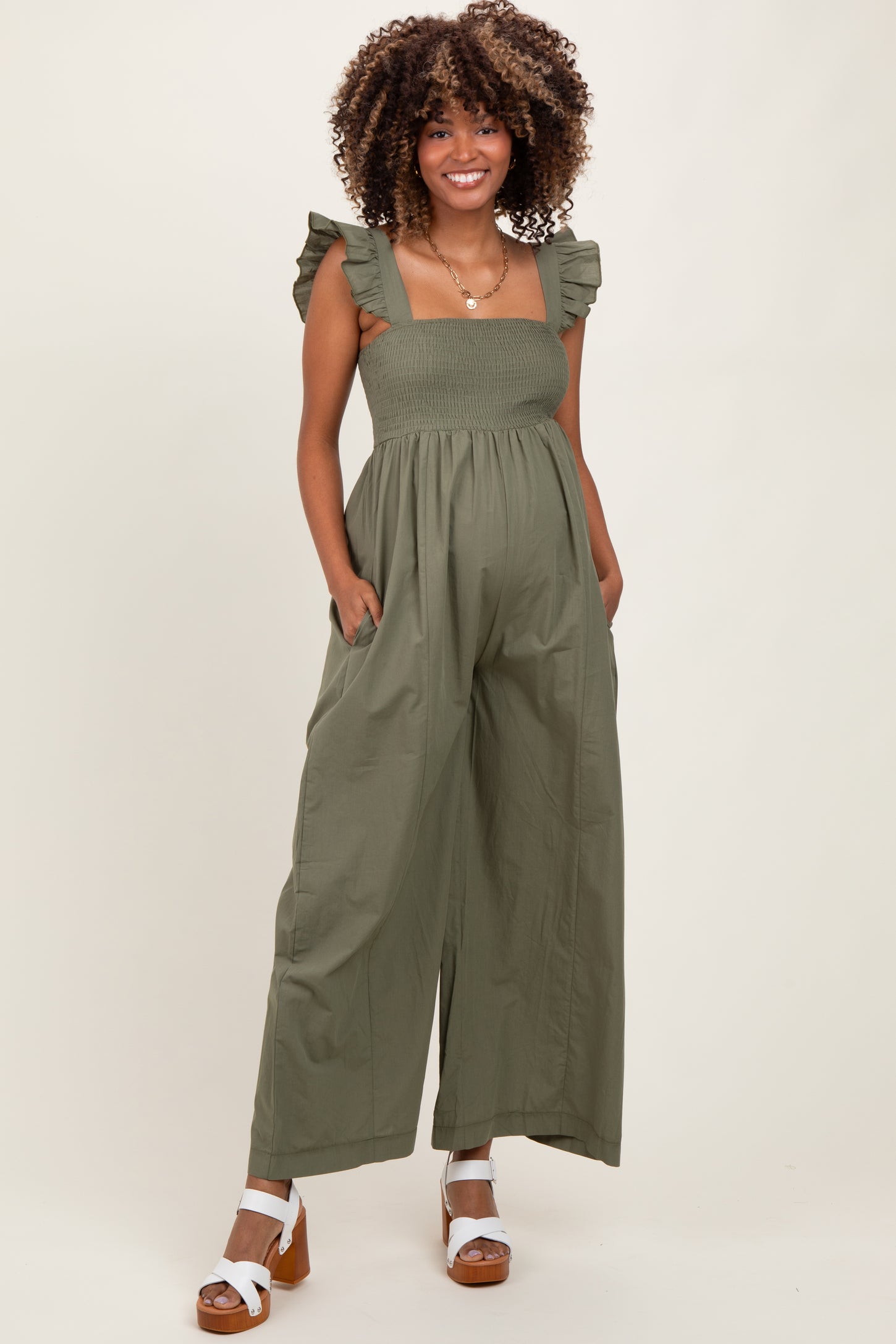 woman wearing a green maternity jumpsuit with ruffled straps and a smocked bodice, showcasing her baby bump