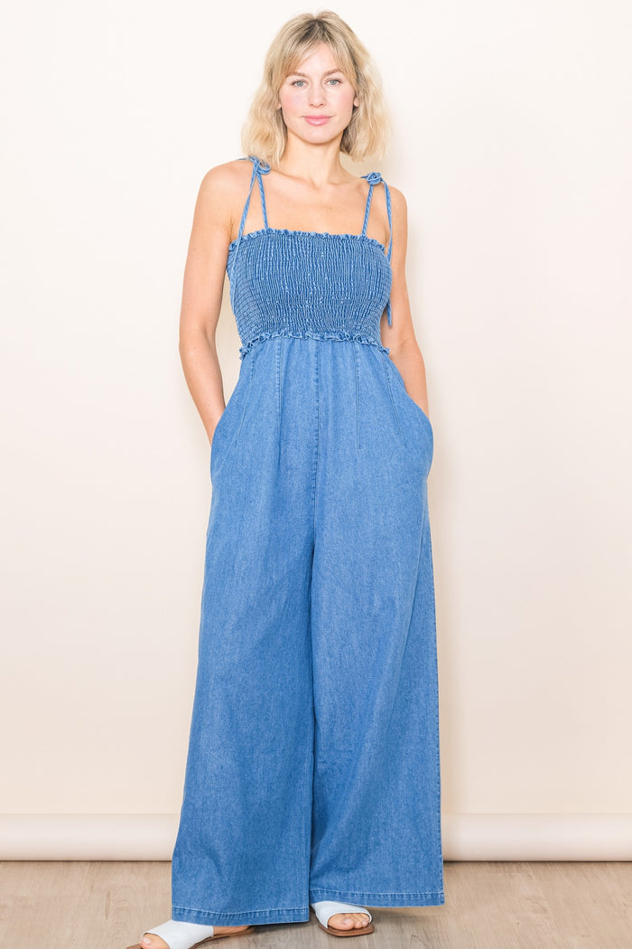 woman wearing a light blue denim jumpsuit with smocked bodice and wide legs, standing with hands in pockets