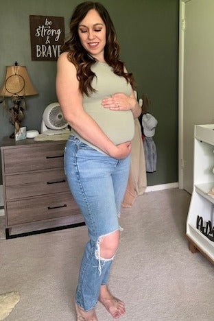 woman wearing a light green maternity tank top and distressed blue jeans, gently cradling her baby bump