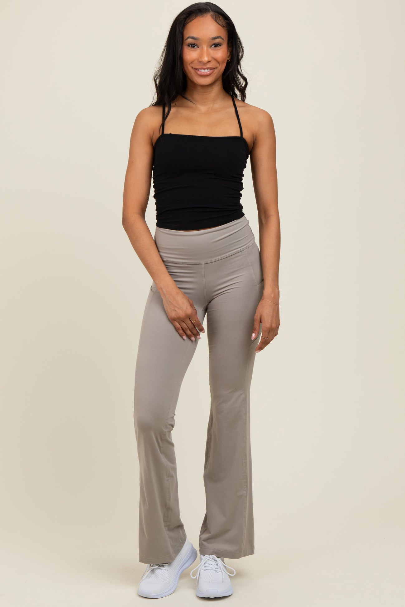 woman wearing a black spaghetti strap top and beige flared yoga pants, standing and smiling