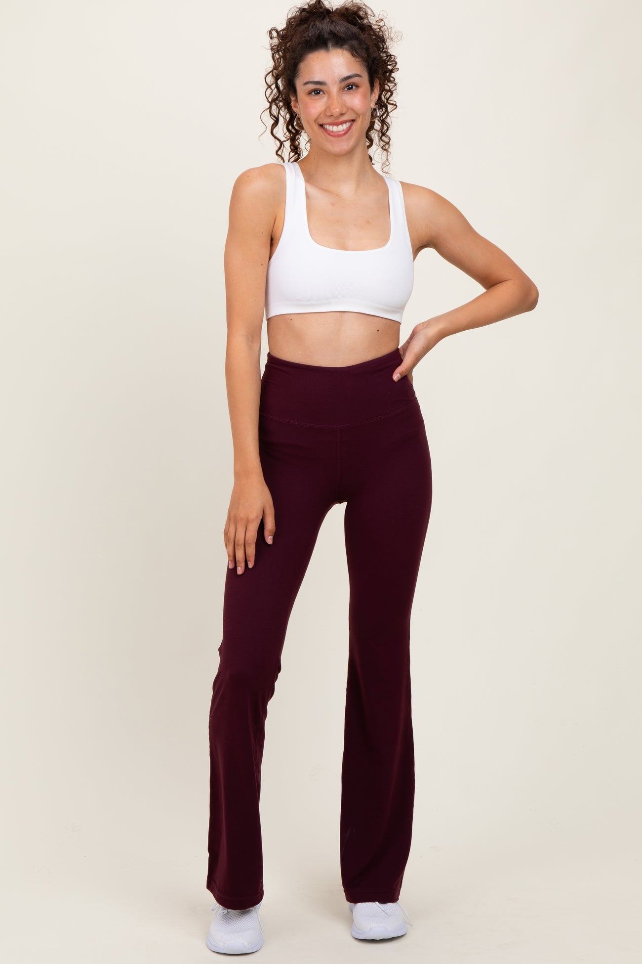 woman wearing a white crop top and burgundy flared workout leggings, smiling and posing with one hand on her hip