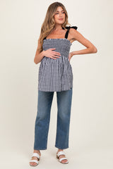 woman wearing a navy and white gingham maternity top with bow detail and holding her baby bump, paired with light blue jeans