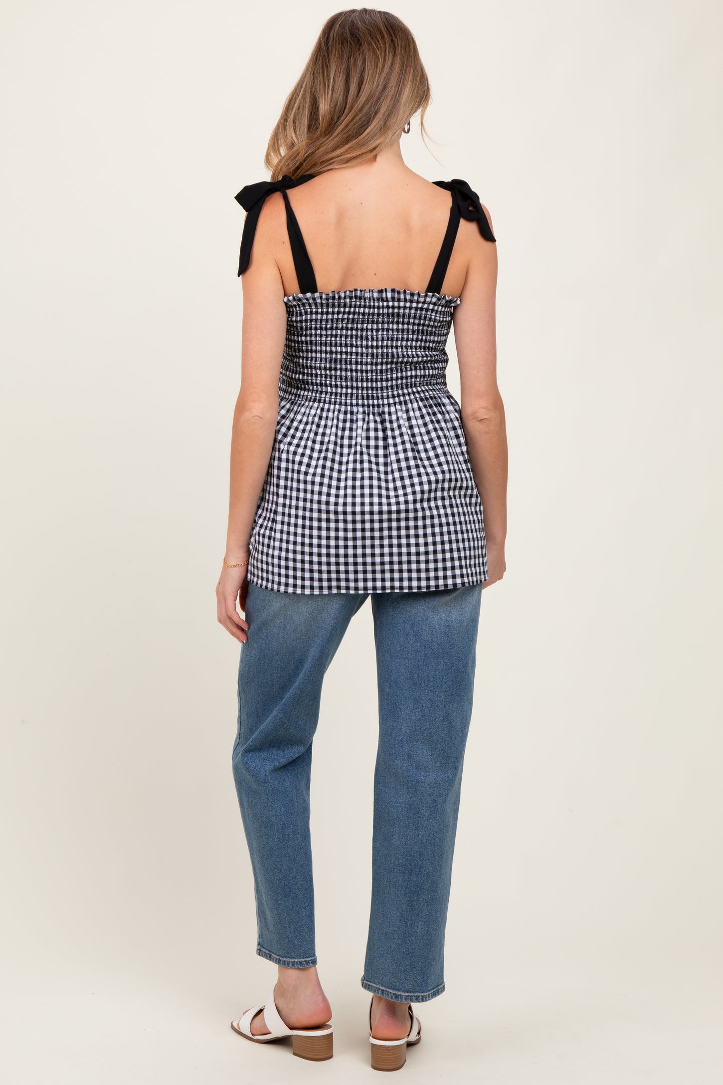 woman wearing a black and white gingham maternity top with adjustable straps and a cinched waist, paired with light blue jeans