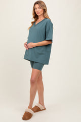 woman wearing a teal maternity top and matching shorts, holding her baby bump while standing on a light background