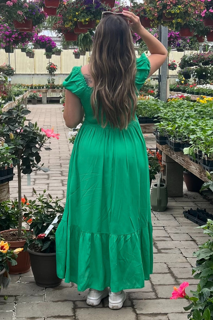woman wearing a green off-the-shoulder maxi dress standing among potted plants in a garden center
