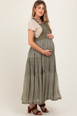 woman wearing a green maternity dress with floral embroidery and a tiered skirt, holding her baby bump