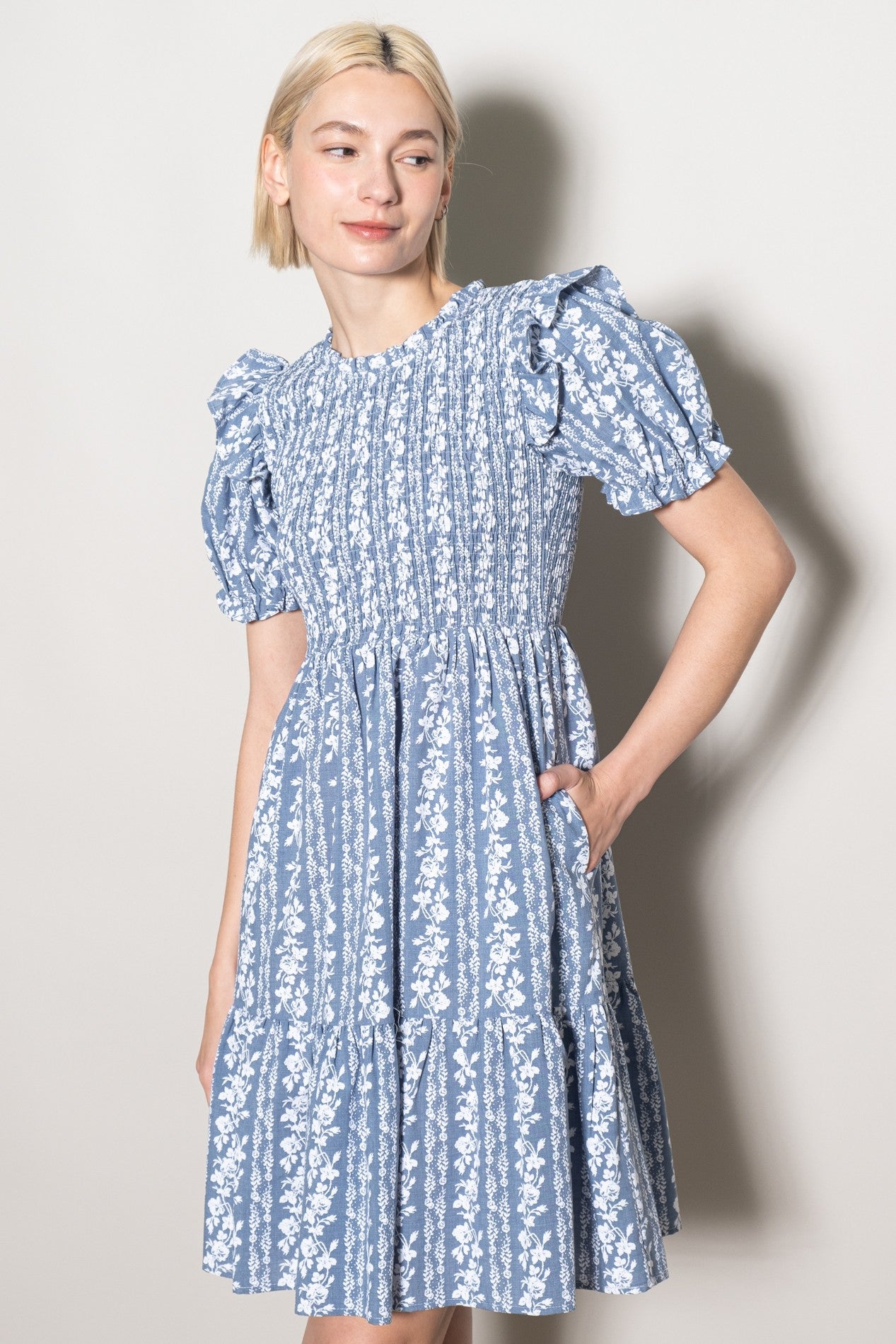 woman wearing a blue floral print short dress with puff sleeves and a tiered skirt, posing with one hand in a pocket