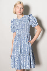 woman wearing a blue floral print short dress with puff sleeves and a tiered skirt, posing with one hand in a pocket