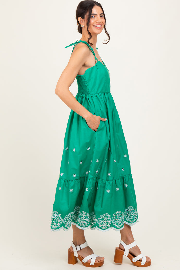 woman wearing a green maxi dress with embroidered details and tie straps, standing with pockets and white chunky heeled sandals