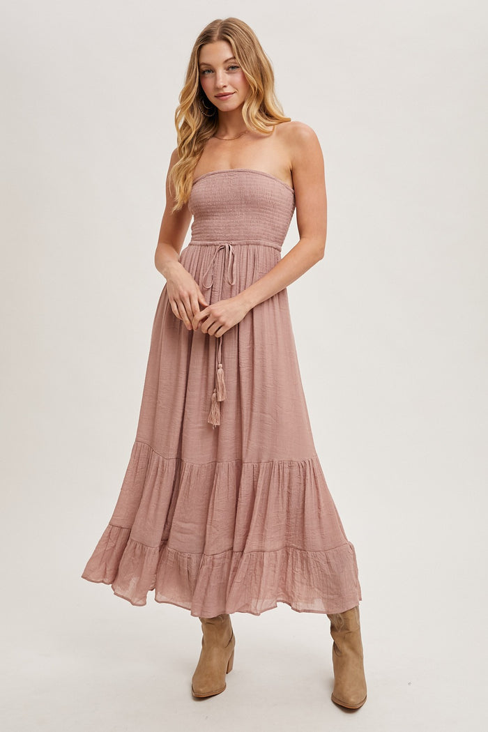 woman wearing a dusty pink strapless maxi dress with a smocked bodice and tiered hem, paired with tan ankle boots