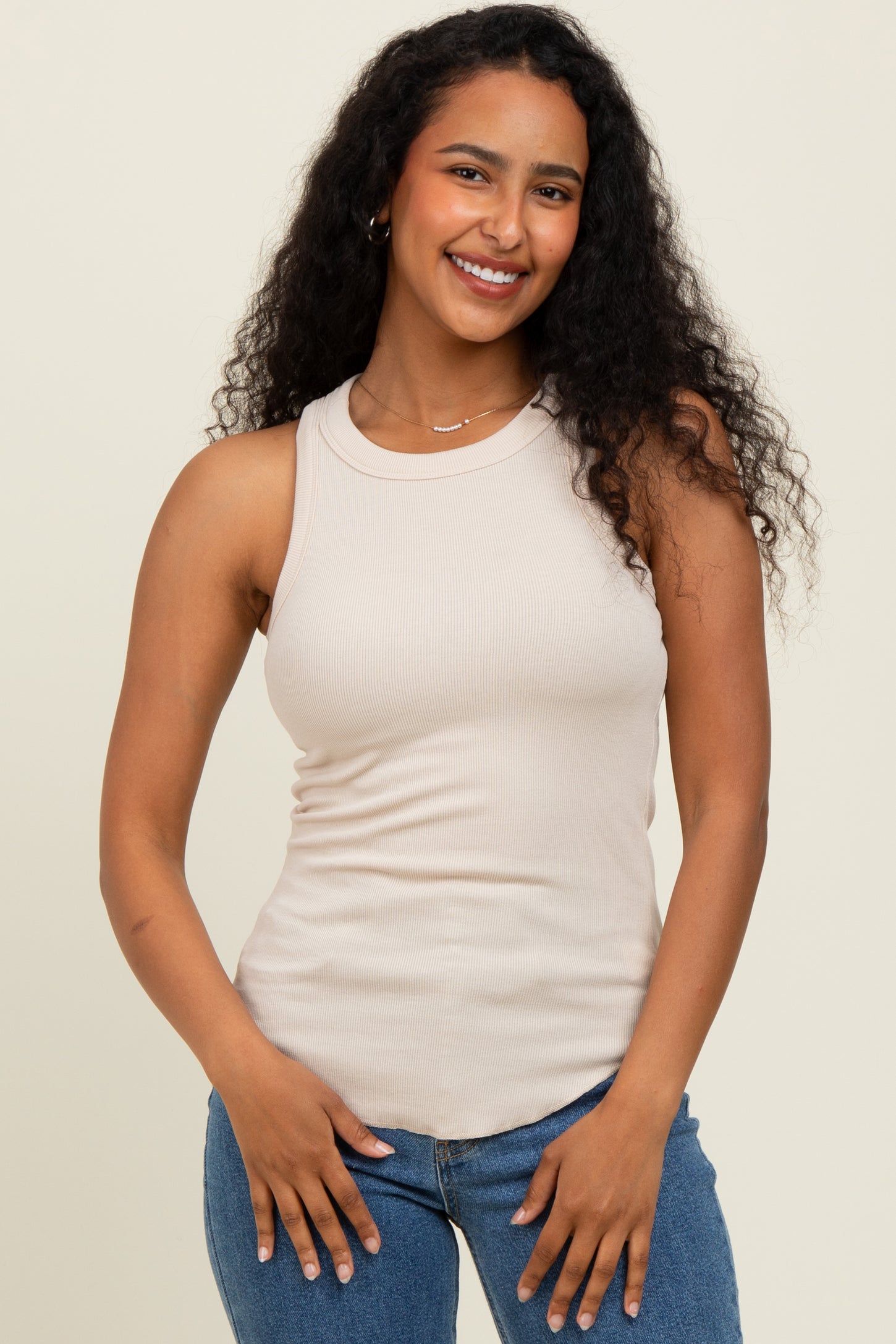 woman wearing a beige ribbed tank top and blue jeans, smiling with curly hair down