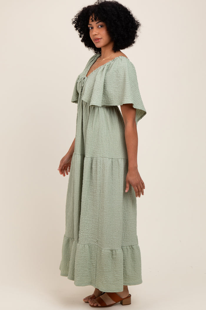 woman wearing a light green tiered maxi dress with a ruffled cape sleeve and a tie detail at the neckline