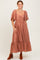 woman wearing a rust-colored maxi dress with puff sleeves and a ruffled hem, posing with hands in pockets