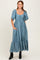 woman wearing a teal maxi dress with short puff sleeves and a tiered skirt, standing with hands in pockets