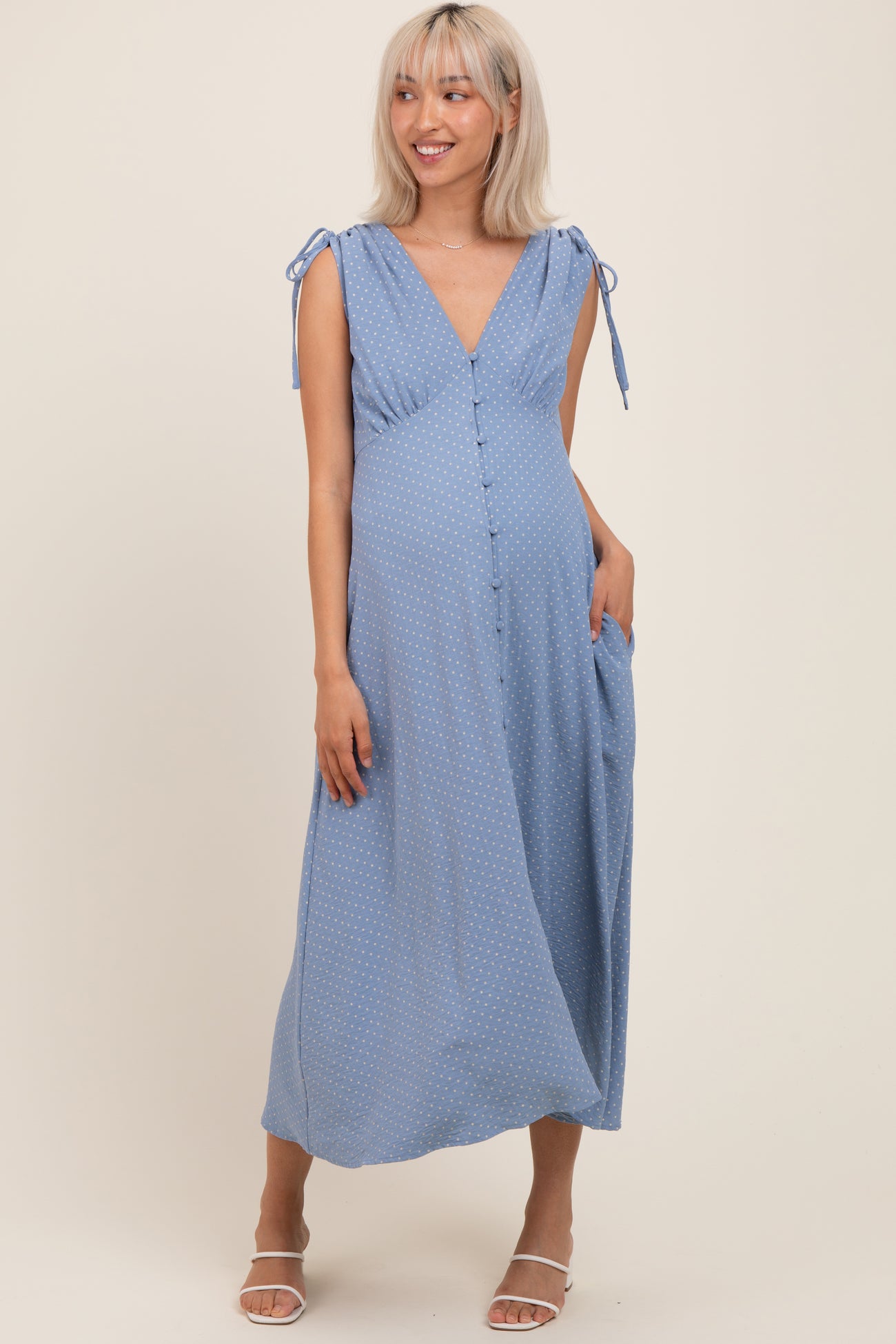 woman wearing a light blue maternity dress with tie straps, button details, and pockets, showcasing her baby bump