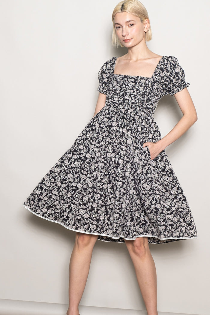 woman wearing a black floral puff sleeve dress with a square neckline and pockets, standing against a neutral background