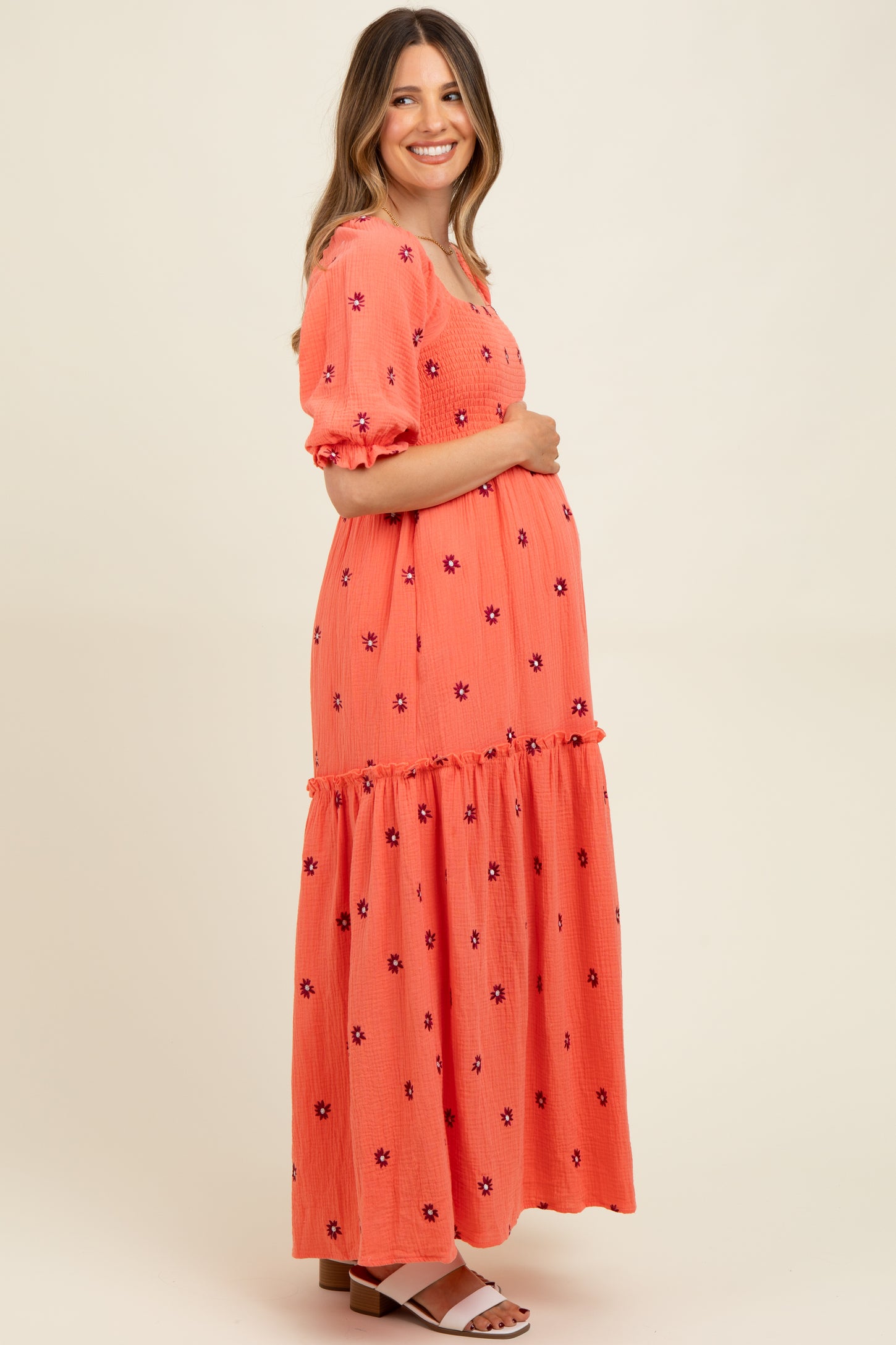 woman wearing a coral floral embroidered maternity maxi dress and holding her baby bump