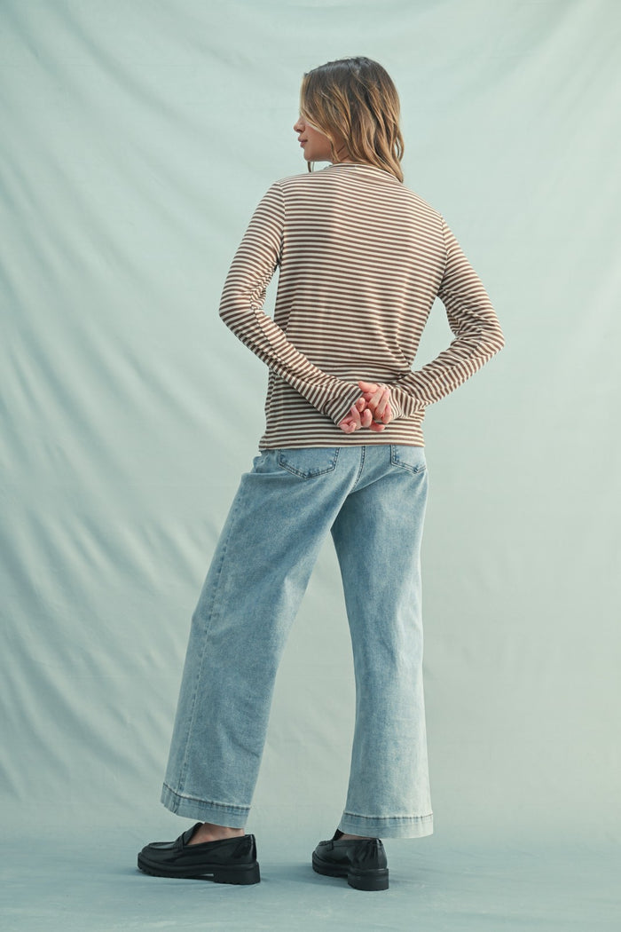 woman wearing a brown and beige striped long sleeve top and light blue wide-leg jeans, standing with hands behind her back