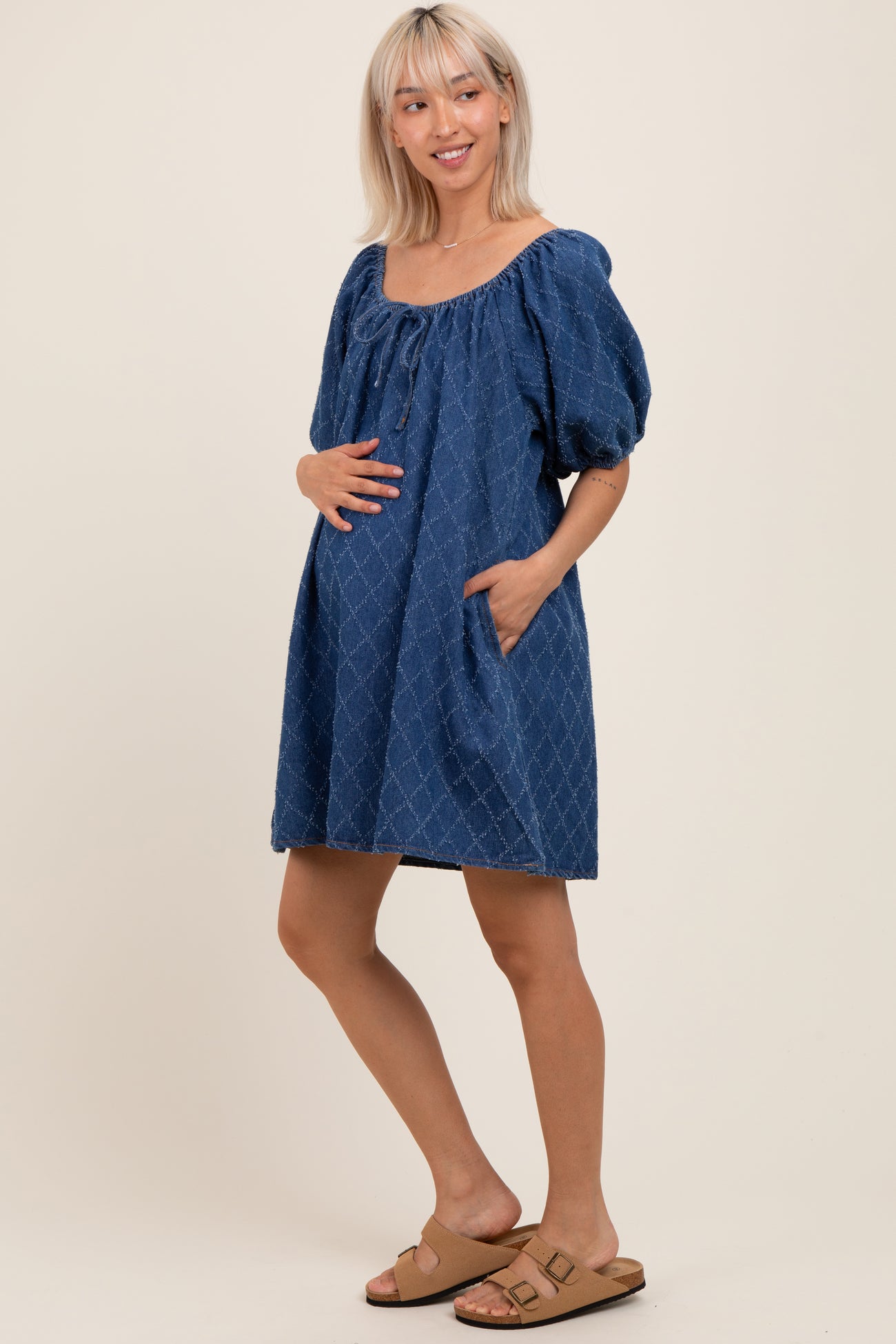 woman wearing a blue maternity dress with puff sleeves and pockets, gently holding her baby bump
