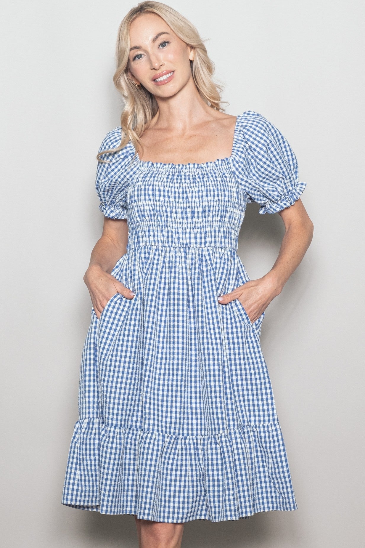 woman wearing a blue and white gingham puff sleeve dress with a smocked bodice and ruffled hem, posing with hands in pockets